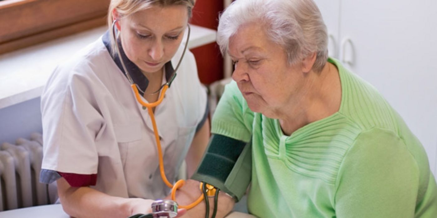 Alter Frau wird von junger Altenpflegerin der Blutdruck mithilfe von Oberarmmanschette und Stethoskop gemessen