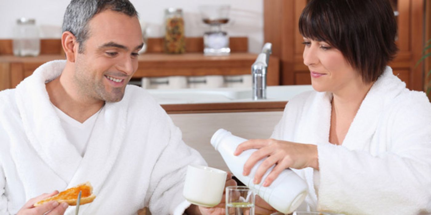 Mann und Frau im mittleren Alter und im Bademantel frühstücken, sie schenkt ihm eine Tasse Milch ein