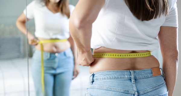 Frau, hält sich ein Maßband an die Taille und schaut in den Spiegel.