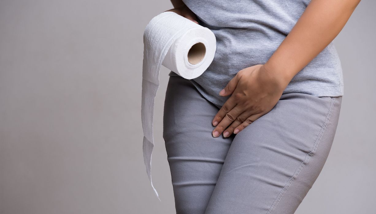 Frau, hält eine Rolle mit Klopapier in der Hand und eine Hand auf ihre Blase.