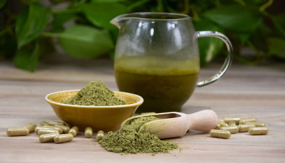 Kratompulver, -kapseln und -tee, im Hintergrund unscharf: Blätter des Kratombaums.