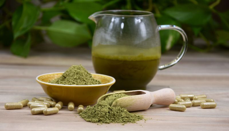 Kratompulver, -kapseln und -tee, im Hintergrund unscharf: Blätter des Kratombaums.