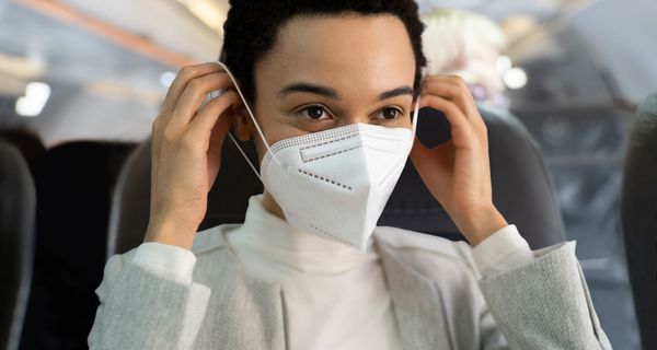 Eine dunkelhäutige Frau mit kurzen Haaren setzt im Flugzeug eine FFP2-Maske auf.