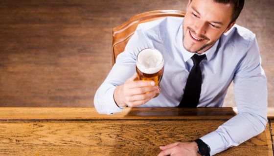 Mann in den 30ern, hellblaues Hemd, Schlips, etwas von oben fotografiert, am Tisch sitzend und ein Feierabendbier in der rechten Hand, auf das er entspannt lächelnd blickt.