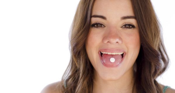 Frontalfoto: junge Frau, dunkle, glatte, lange Haare, zeigt Zunge, auf der eine Tablette liegt, in die Kamera