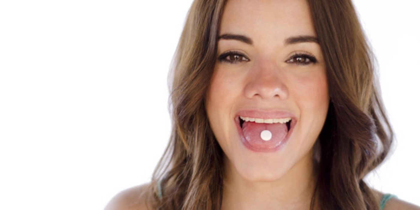 Frontalfoto: junge Frau, dunkle, glatte, lange Haare, zeigt Zunge, auf der eine Tablette liegt, in die Kamera