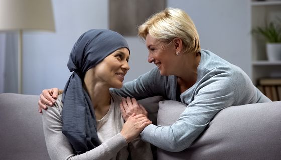 Krebspatientin, wird von hinten von einer älteren Frau umarmt, beide lächeln.