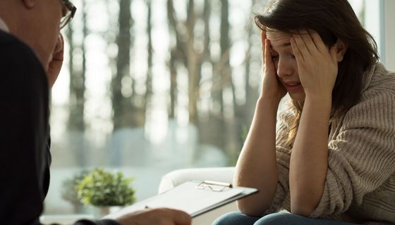 Psychotherapie-Sitzung: Eine belastete Frau berichtet, beide Hände in ihrem Gesicht, die Fingerkuppen an den Schläfen. Ihr gegenüber sitzt ein älterer Herr mit einem Klemmbrett.