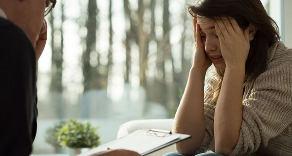 Psychotherapie-Sitzung: Eine belastete Frau berichtet, beide Hände in ihrem Gesicht, die Fingerkuppen an den Schläfen. Ihr gegenüber sitzt ein älterer Herr mit einem Klemmbrett.