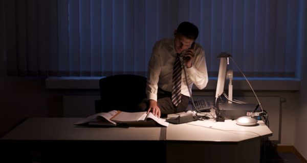 Mann arbeitet spät in der Nacht in dunklem Büro, nur die Schreibtischlampe brennt