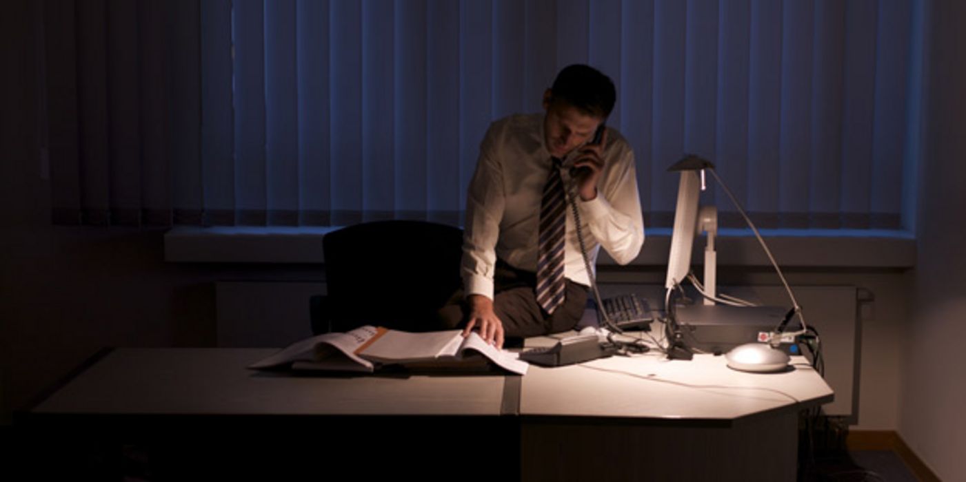 Mann arbeitet spät in der Nacht in dunklem Büro, nur die Schreibtischlampe brennt