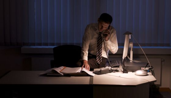 Mann arbeitet spät in der Nacht in dunklem Büro, nur die Schreibtischlampe brennt