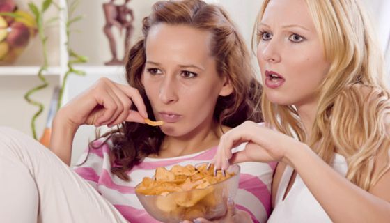 Zwei junge Frauen sitzen zusammen auf der Couch, schauen Fernsehen und essen Chips.