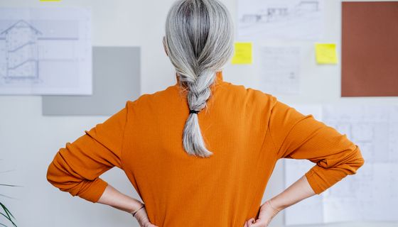 Frau mit grauen Haaren, steht mit dem Gesicht zu einer Wand.