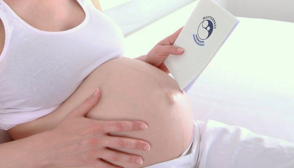 Profilbild einer Schwangeren: ab Halsansatz bis Beinansatz, weißes Top, bauchfrei, rechte Hand am Bauch, linke Hand hält einen Mutterpass