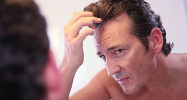 Porträtfoto: Mann, in den frühen 40ern, betrachtet seine Geheimratsecken (dunkle, zurückgekämmte Haare) in einem Spiegel und fasst dabei mit einer Hand ins Haar