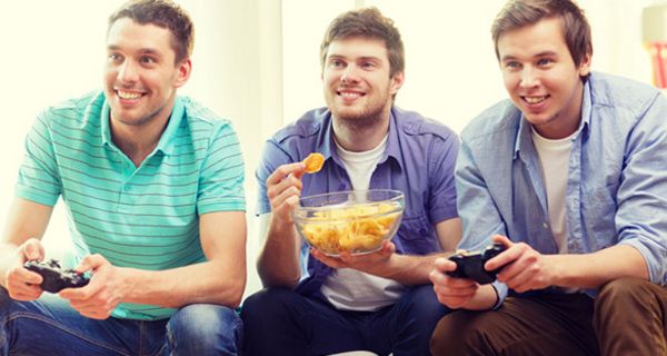 3 junge Männer auf Couch, 2 mit Playstation (re. u. li.), 1 mit Kartoffelchips in einer Schüssel