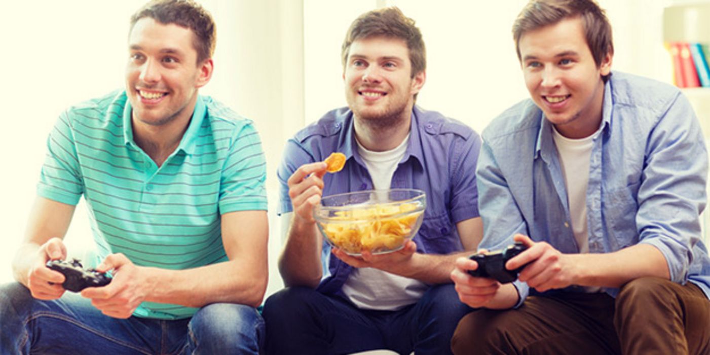 3 junge Männer auf Couch, 2 mit Playstation (re. u. li.), 1 mit Kartoffelchips in einer Schüssel