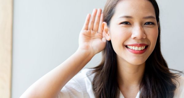 Junge Frau, hält eine Hand hinter ihr Ohr und lächelt in die Kamera.