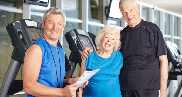 Szene im Fitness-Studio: Trainer und Seniorenpaar lachen in die Kamera, Mann hat Arm um die Frau gelegt