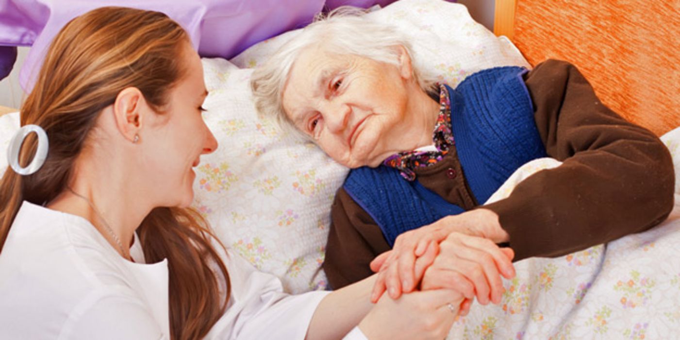 Junge Pflegerin hält die Hand einer alten, pflegebedürftigen Frau, die im Bett liegt