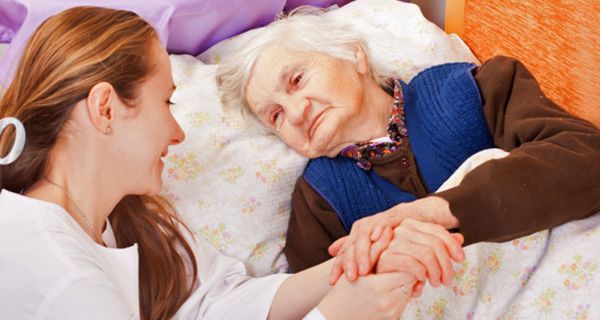 Junge Pflegerin hält die Hand einer alten, pflegebedürftigen Frau, die im Bett liegt