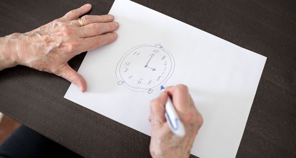 Eine alte Frauenenhand zeichnet ein Ziffernblatt mit falschen Zahlen auf ein Blatt Papier.