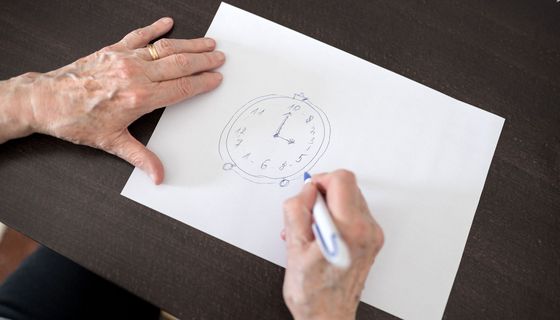 Eine alte Frauenenhand zeichnet ein Ziffernblatt mit falschen Zahlen auf ein Blatt Papier.