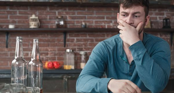 Ein Mann blickt nachdenklich ins Leere, neben ihm stehen zwei fast leere Flaschen, eine liegt umgekippt daneben.