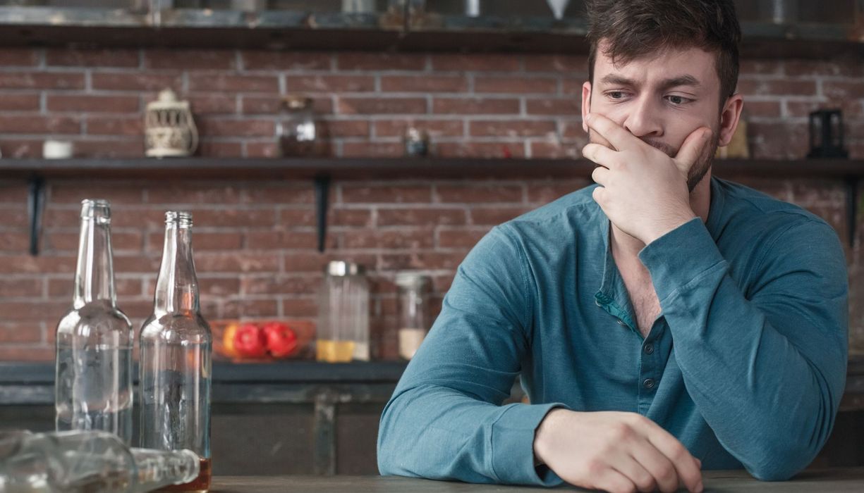 Ein Mann blickt nachdenklich ins Leere, neben ihm stehen zwei fast leere Flaschen, eine liegt umgekippt daneben.