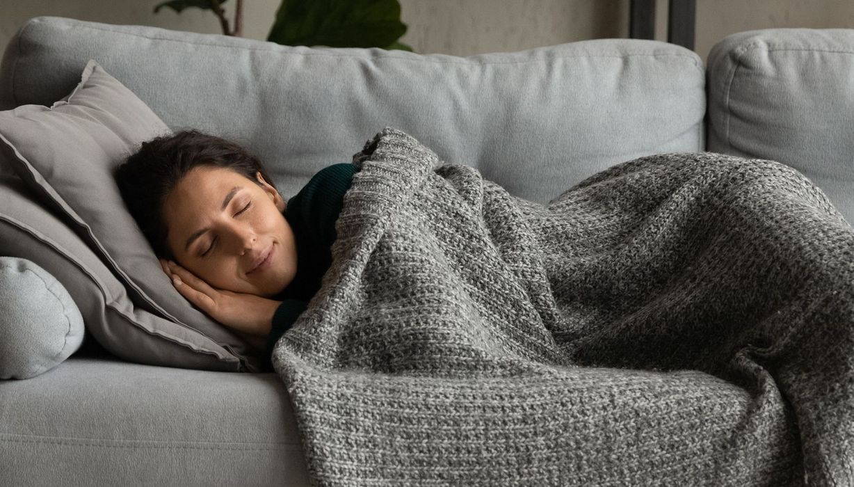 Eine Frau schläft bei Tag auf einem Sofa. Sie lächelt selig.