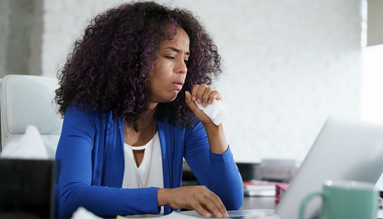 Junge Frau in Bürokleidung hustet mit einem Taschentuch in der Hand, während sie am Laptop arbeitet.