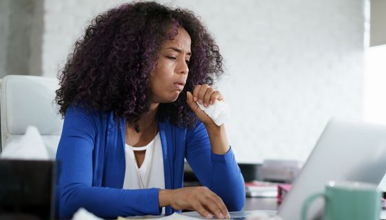 Junge Frau in Bürokleidung hustet mit einem Taschentuch in der Hand, während sie am Laptop arbeitet.