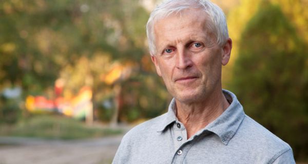 Portraitfoto (Brustbild): Mann (ca. Mitte/Ende 60), schlank, grauhaarig, graues Poloshirt, freundlich in die Kamera blickend, Hintergrund schemenhaft Parklandschaft