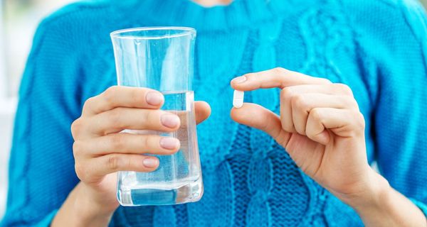 Frau mit blauem Pullover hält ein Glas Wasser und eine Tablette in den Händen.