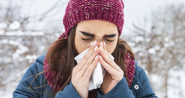 Junge Frau mit Mütze und Winterjacke niest in einem verschneiten Wäldchen.
