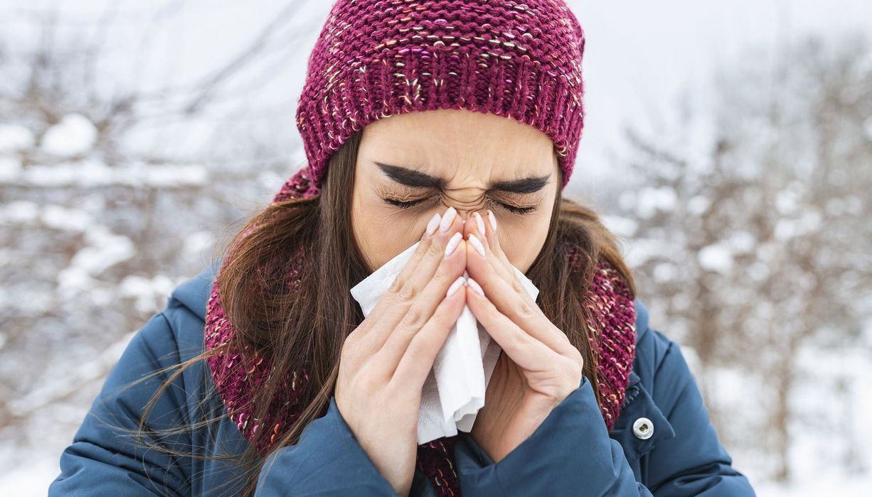 Junge Frau mit Mütze und Winterjacke niest in einem verschneiten Wäldchen.