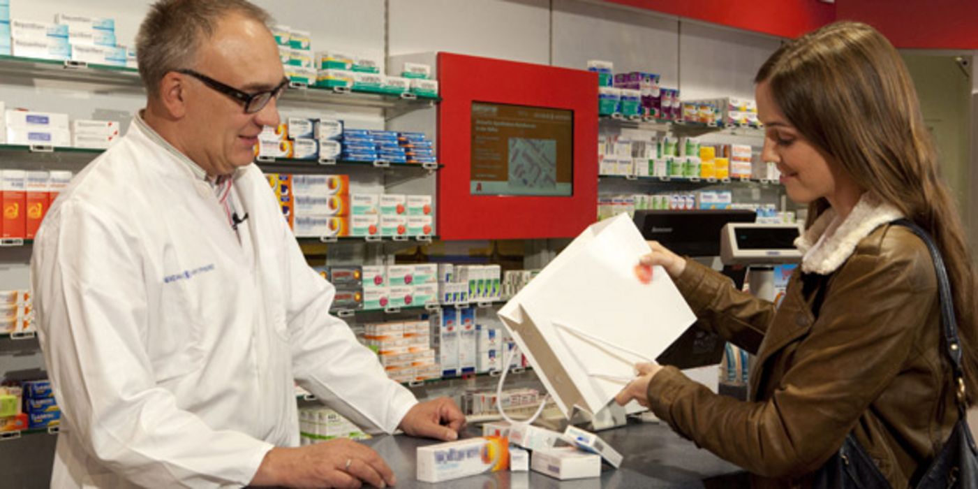 Apotheker mit Kundin, die eine Tüte mit Medikamenten von ihm in Augenschein nehmen lässt