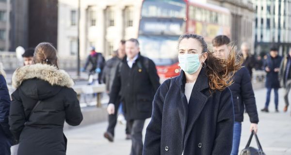 Junge Frau mit Maske, läuft in London auf einem Fußgängerweg, Doppeldecker-Bus im Hintergrund.