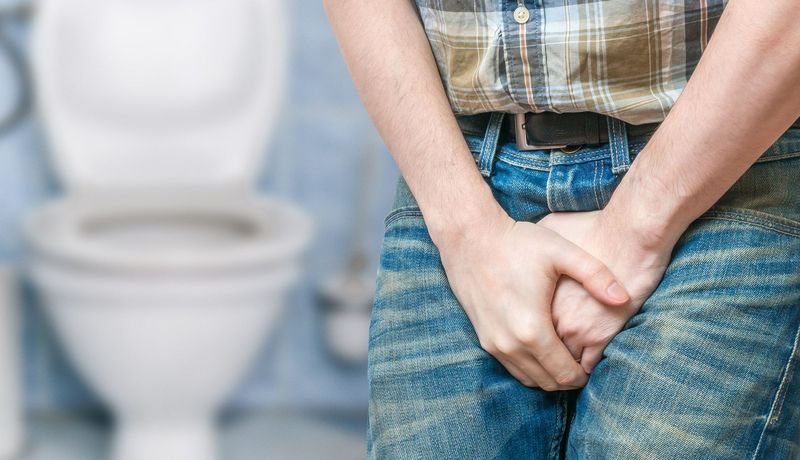 Vordergrund: Bekleideter Mann hält sich den Schritt, Hintergrund (unscharf): Eine Toilette.