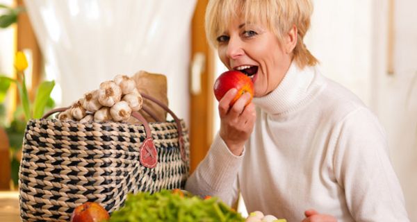 Blonde Frau um die 50 beißt in einen Apfel, auf dem Tisch grüner Salat, Apfel, Möhre und ein Korb, aus dem Knoblauchstränge herausschauen