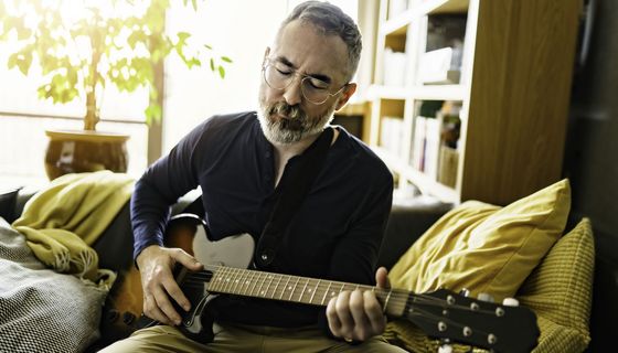 Älterer Mann mit Brille spielt auf seiner Couch mit geschlossenen Augen Gitarre.