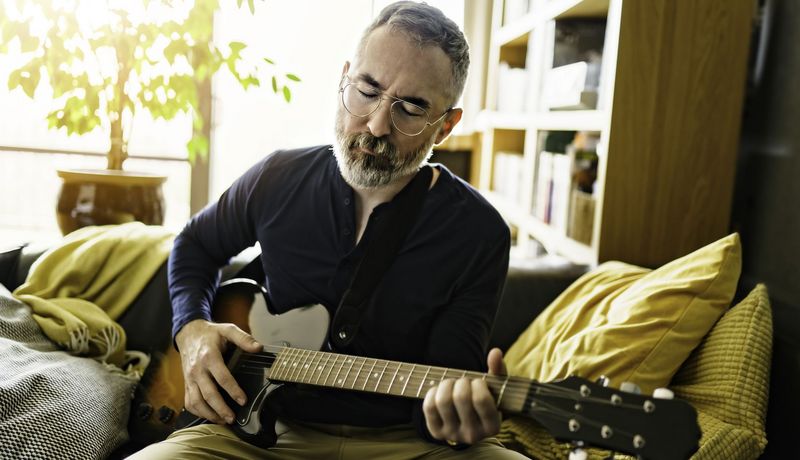 Älterer Mann mit Brille spielt auf seiner Couch mit geschlossenen Augen Gitarre.