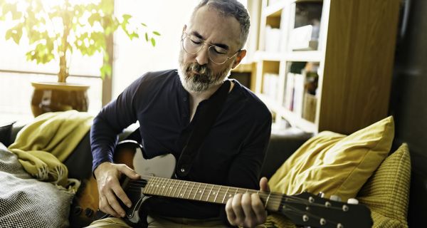 Älterer Mann mit Brille spielt auf seiner Couch mit geschlossenen Augen Gitarre.
