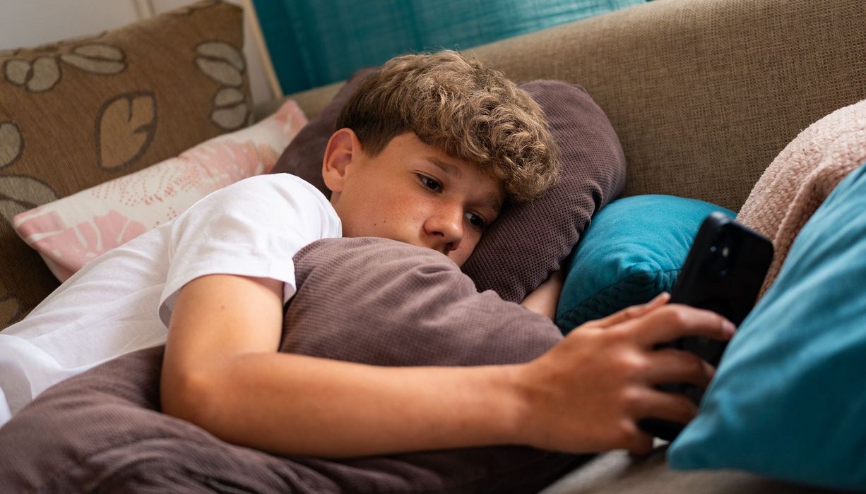 Unglücklicher Jugendlicher liegt auf einem Sofa, das Handy in der Hand.
