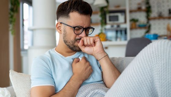 Ein junger Mann sitzt auf dem Sofa, hält sich eine Hand vor dem Mund und die andere vor die Brust.