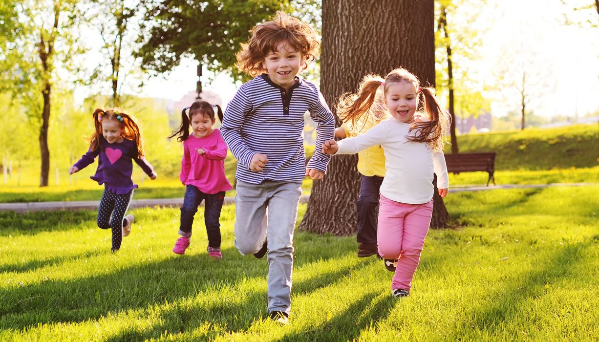 Fünf Kinder rennen über eine Wiese im Schatten eines Baumes.
