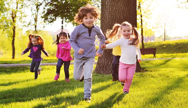 Fünf Kinder rennen über eine Wiese im Schatten eines Baumes.
