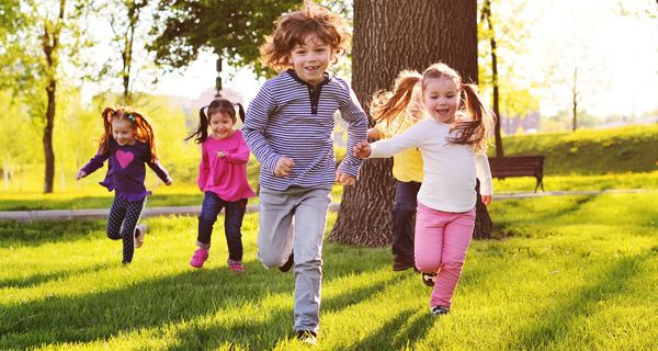 Fünf Kinder rennen über eine Wiese im Schatten eines Baumes.