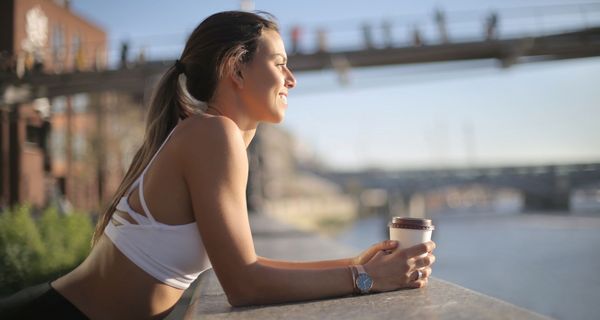 Junge Frau, lehnt sich mit einem Kaffee an eine Brüstung, sportliches Outfit.